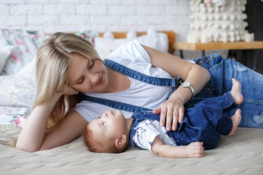 Yeni doğmuş çocuğunu tutan genç anne. Kadın ve yeni doğan çocuk beyaz bir yatak odasında rahatlayın. Genç bir anne kucağında yeni doğan oğlunu nazikçe tutar. Evde aile. Yeni doğan oğlunu whith oynayan genç anne