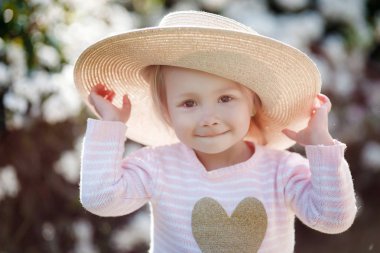 Büyük hasır şapkalı sevimli bir kız, beyaz çiçekli çalıların arka planına bakarak, bir bahar parkı parkında tek başına vakit geçirir. Çiçekli bir parkta yürüyen sevimli bir küçük kızın bahar portresi.