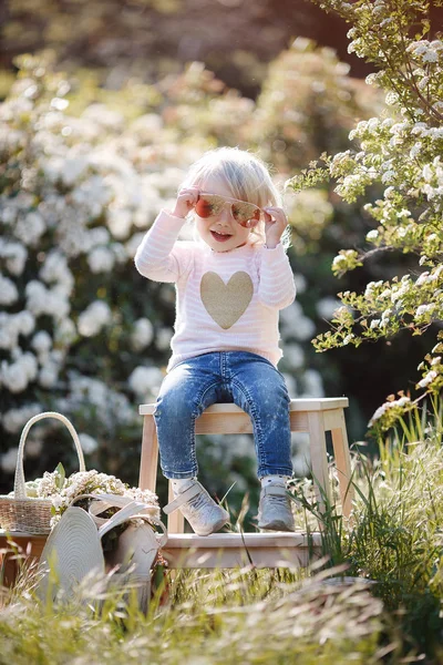 大きなわら帽子をかぶったかわいい女の子は 白い開花茂みを背景に座って 春の公園で一人で屋外で時間を過ごします 花の咲く公園を歩く魅力的な少女の春の肖像画 — ストック写真