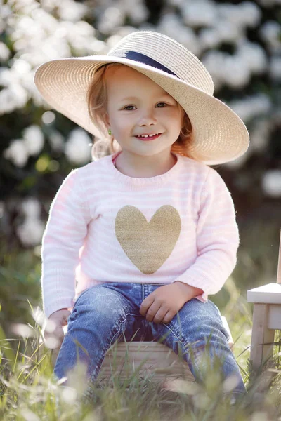 大きなわら帽子をかぶったかわいい女の子は 白い開花茂みを背景に座って 春の公園で一人で屋外で時間を過ごします 花の咲く公園を歩く魅力的な少女の春の肖像画 — ストック写真