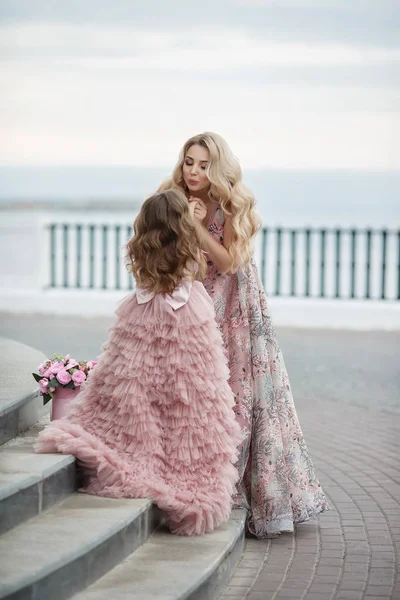 美しいスレンダー女性と彼女の小さな娘は 春に屋外で一緒に楽しんで 開花桃と宮殿の路地に ピンク色の美しいボールガウンに身を包んだ 路上で美しいイブニングドレスでママと娘 — ストック写真