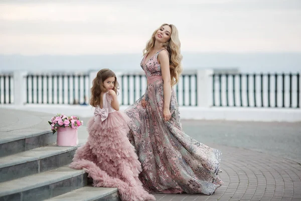 美しいスレンダー女性と彼女の小さな娘は 春に屋外で一緒に楽しんで 開花桃と宮殿の路地に ピンク色の美しいボールガウンに身を包んだ 路上で美しいイブニングドレスでママと娘 — ストック写真