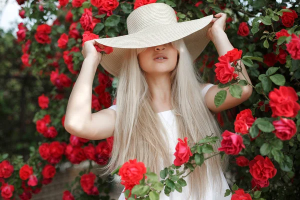 香水和化妆品 女人在盛开的玫瑰花丛前 野玫瑰的花 玫瑰的香气 女孩可爱的金发嗅香粉红色绽放 年轻的美丽的女人在帽子 附近的一大丛红玫瑰在春天的花园户外 — 图库照片