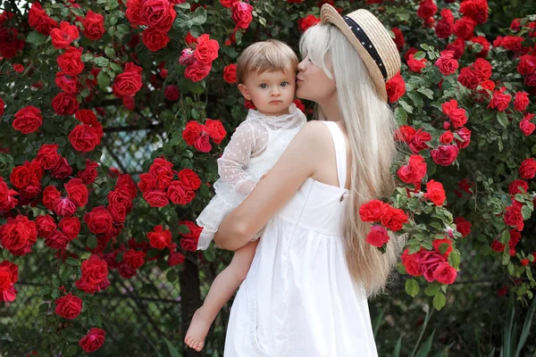 一起快乐的家庭时光 年轻的母亲带着一个小女儿在公园里 在一棵大灌木丛旁 盛开的红玫瑰 快乐的男女与美丽的春天的花朵在绿色的背景下 家庭度假概念 母亲节 — 图库照片