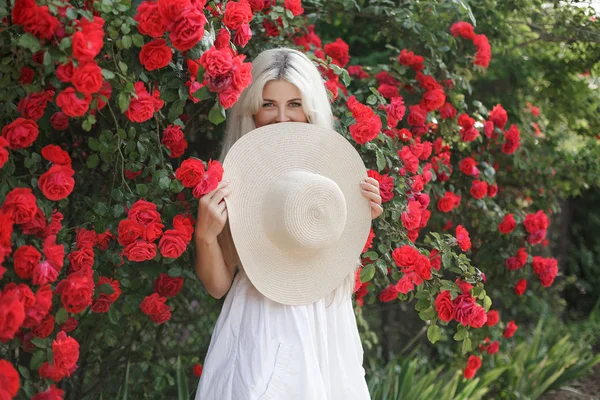 Perfume and cosmetics. Woman in front of blooming roses bush. Blossom of wild roses.  Aroma of roses. Girl adorable blonde sniffing fragrance of pink bloom. Young beautiful woman in a hat, near a large bush of red roses in the spring garden outdoors