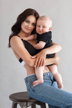 Denim tulumgiyen genç kadın anne kucağında bir bebek tutuyor. Stüdyoda beyaz arka plan. Bebeği beyaza izole edilmiş bir anne. Anneler Günü, aşk ailesi, ebeveynlik çocukluk kavramı. Yeni doğmuş mutlu bir aile. Portret anne ve küçük oğlu.