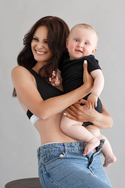 デニムのオーバーオールを着た若い女性の母親は 赤ちゃんの子供を腕に抱いている スタジオの白い背景 赤ちゃんを持つ母親は白で孤立しています 母の日 愛の家族 親の子供時代の概念 新生児と幸せな家族 ポートレットの母と小さな息子 — ストック写真