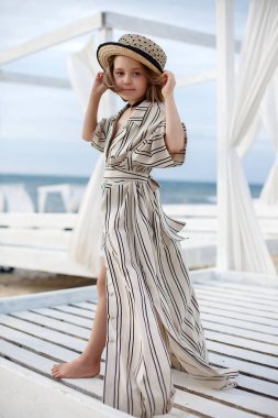 Küçük (8 yaşında) beyaz çizgili elbise ve pembe güneş gözlüğü içinde oldukça neşeli kız ahşap beyaz bir iskele üzerinde duruyor.8 yaşındaki kız sıcak güneşli bir günde tek başına tropikal plajda istirahat. Yaz tatillerinde tropik bir plajda poz veren çocuk