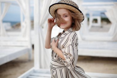 Küçük (8 yaşında) beyaz çizgili elbise ve pembe güneş gözlüğü içinde oldukça neşeli kız ahşap beyaz bir iskele üzerinde duruyor.8 yaşındaki kız sıcak güneşli bir günde tek başına tropikal plajda istirahat. Yaz tatillerinde tropik bir plajda poz veren çocuk