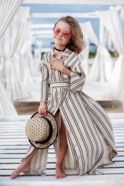 Küçük (8 yaşında) beyaz çizgili elbise ve pembe güneş gözlüğü içinde oldukça neşeli kız ahşap beyaz bir iskele üzerinde duruyor.8 yaşındaki kız sıcak güneşli bir günde tek başına tropikal plajda istirahat. Yaz tatillerinde tropik bir plajda poz veren çocuk