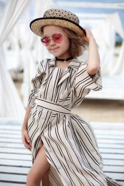 Küçük (8 yaşında) beyaz çizgili elbise ve pembe güneş gözlüğü içinde oldukça neşeli kız ahşap beyaz bir iskele üzerinde duruyor.8 yaşındaki kız sıcak güneşli bir günde tek başına tropikal plajda istirahat. Yaz tatillerinde tropik bir plajda poz veren çocuk