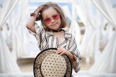 Küçük (8 yaşında) beyaz çizgili elbise ve pembe güneş gözlüğü içinde oldukça neşeli kız ahşap beyaz bir iskele üzerinde duruyor.8 yaşındaki kız sıcak güneşli bir günde tek başına tropikal plajda istirahat. Yaz tatillerinde tropik bir plajda poz veren çocuk