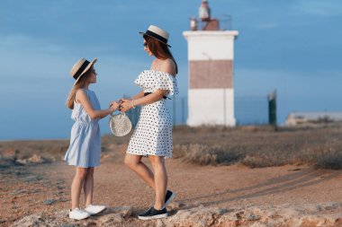 Neşeli bir çocuk ve annesi su kenarında güneşli bir sabah tadını çıkarıyor. Güzel bir esmer anne ve kızı nın portresi arka planda deniz ile kayalık bir sahilde güzel elbiseler içinde. Mutlu aile tatil tatil geçirir.
