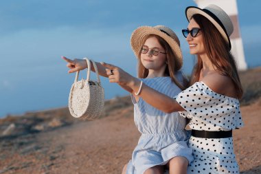 Neşeli bir çocuk ve annesi su kenarında güneşli bir sabah tadını çıkarıyor. Güzel bir esmer anne ve kızı nın portresi arka planda deniz ile kayalık bir sahilde güzel elbiseler içinde. Mutlu aile tatil tatil geçirir.