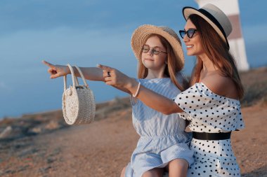 Neşeli bir çocuk ve annesi su kenarında güneşli bir sabah tadını çıkarıyor. Güzel bir esmer anne ve kızı nın portresi arka planda deniz ile kayalık bir sahilde güzel elbiseler içinde. Mutlu aile tatil tatil geçirir.