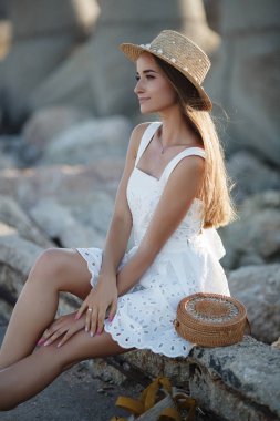 Güzel esmer kayalar üzerinde oturan. Seksi esmer kız akşam plajda tek başına poz. Genç oldukça sıcak seksi çekici kız arka planda mavi deniz ve gökyüzü ile taşlar üzerinde beyaz bir elbise ve şapka içinde rahatlatıcı. Yaz Tatili Konsepti.