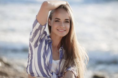 Güzel esmer kayalar üzerinde oturan. Seksi esmer kız akşam plajda tek başına poz. Genç oldukça sıcak seksi çekici kız çizgili bir gömlek ve arka planda mavi deniz ve gökyüzü ile taşlar üzerinde şort rahatlatıcı. Yaz Tatili Konsepti.