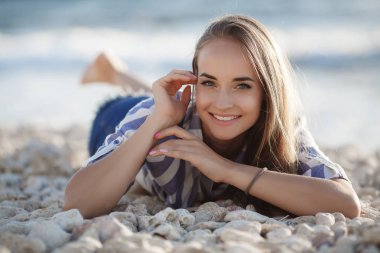 Güzel esmer kayalar üzerinde oturan. Seksi esmer kız akşam plajda tek başına poz. Genç oldukça sıcak seksi çekici kız çizgili bir gömlek ve arka planda mavi deniz ve gökyüzü ile taşlar üzerinde şort rahatlatıcı. Yaz Tatili Konsepti.