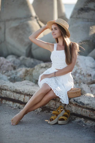 岩の上に座って美しいブルネット セクシーなブルネットの女の子は 夜にビーチで一人でポーズ 白いドレスと青い海と空を背景に石に帽子でリラックス若いかなりホットセクシーな魅力的な女の子 夏休みコンセプト — ストック写真
