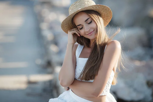 岩の上に座って美しいブルネット セクシーなブルネットの女の子は 夜にビーチで一人でポーズ 白いドレスと青い海と空を背景に石に帽子でリラックス若いかなりホットセクシーな魅力的な女の子 夏休みコンセプト — ストック写真