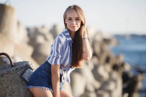 岩の上に座って美しいブルネット セクシーなブルネットの女の子は 夜にビーチで一人でポーズ 青い海と空を背景に石の上にストライプのシャツとショートパンツでリラックス若いかなりホットセクシーな魅力的な女の子 夏休みコンセプト — ストック写真