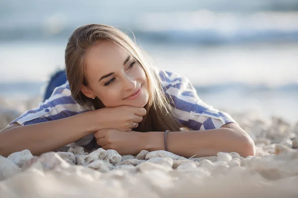 岩の上に座って美しいブルネット セクシーなブルネットの女の子は 夜にビーチで一人でポーズ 青い海と空を背景に石の上にストライプのシャツとショートパンツでリラックス若いかなりホットセクシーな魅力的な女の子 夏休みコンセプト — ストック写真