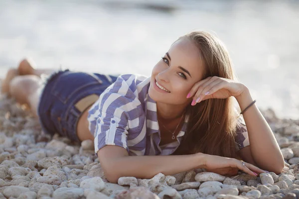 岩の上に座って美しいブルネット セクシーなブルネットの女の子は 夜にビーチで一人でポーズ 青い海と空を背景に石の上にストライプのシャツとショートパンツでリラックス若いかなりホットセクシーな魅力的な女の子 夏休みコンセプト ストック写真