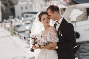 Bir çift aşık, yatların yanındaki iskelede yürüyor. Yazın açık havada aşk yaşayan genç bir çift, arka planda güzel beyaz yatlar ve sakin bir denizde rıhtımda kucaklaşıyorlar. Harika bir çift yeni evli çift, deniz kenarında rıhtımda yürüyor.