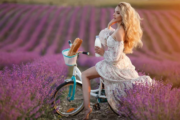 Hübsche junge blonde Frau mit langen Haaren, die auf einem Fahrrad im violetten Lavendelfeld sitzt. Brot und Milch im Korb — Stockfoto