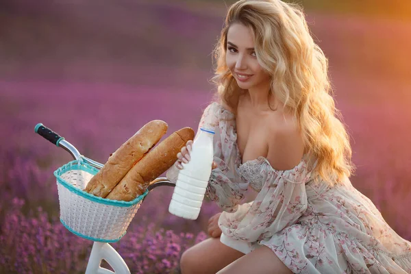 Bonita mujer rubia joven con el pelo largo sentado en bicicleta en el campo de lavanda violeta. pan y leche en cesta — Foto de Stock