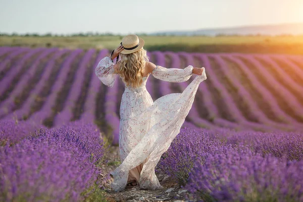 En söt blond kvinna som springer iväg på lavendelfältet. kvinna i lång klänning och halm hatt har kul i blommor av lavendel — Stockfoto