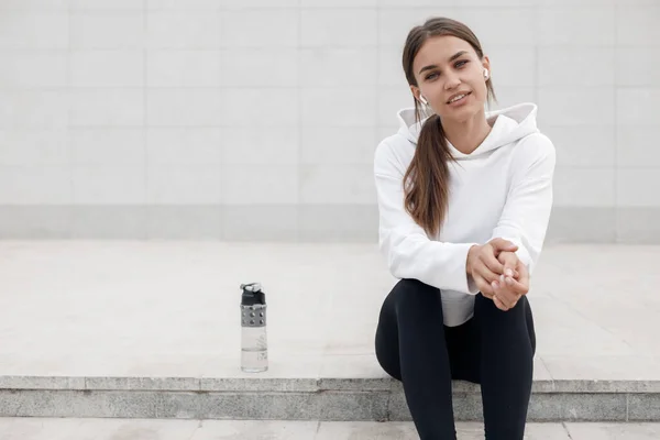 Retrato de una joven en forma y deportiva sentada con una botella de agua al aire libre en la ciudad — Foto de Stock