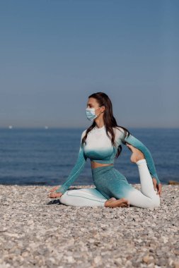 Maskeli genç kadın dışarıda yoga yapıyor.
