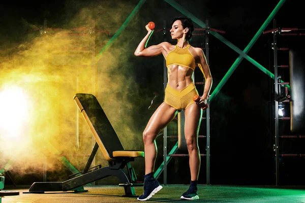 Jovem mulher no ginásio com um halteres, em um belo sportswear, contra um fundo escuro com luzes de cor . — Fotografia de Stock