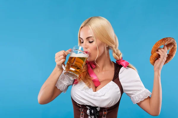 Junges sexy Oktoberfestmädchen - Kellnerin, in traditioneller bayerischer Tracht, serviert große Bierkrüge und isst Brezel auf blauem Hintergrund. Kopierraum. — Stockfoto