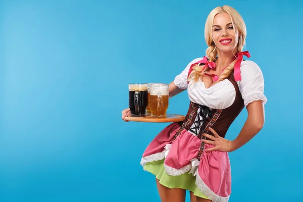Fiatal szexi Oktoberfest lány - pincérnő, egy hagyományos bajor ruhát visel, szolgáló nagy sör bögrék a kék háttér. — Stock Fotó