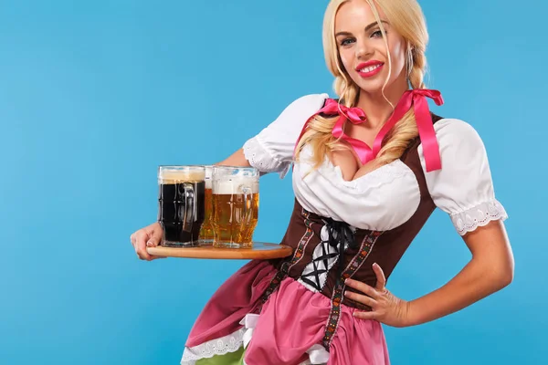 Fiatal szexi Oktoberfest lány - pincérnő, egy hagyományos bajor ruhát visel, szolgáló nagy sör bögrék a kék háttér. — Stock Fotó
