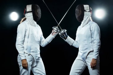 Young fencer athlete wearing mask and white fencing costume. holding the sword on black background with lights. clipart