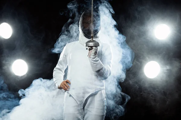 Joven atleta esgrimista con traje de esgrima sosteniendo la espada y la máscara. Aislado sobre fondo negro con luces — Foto de Stock