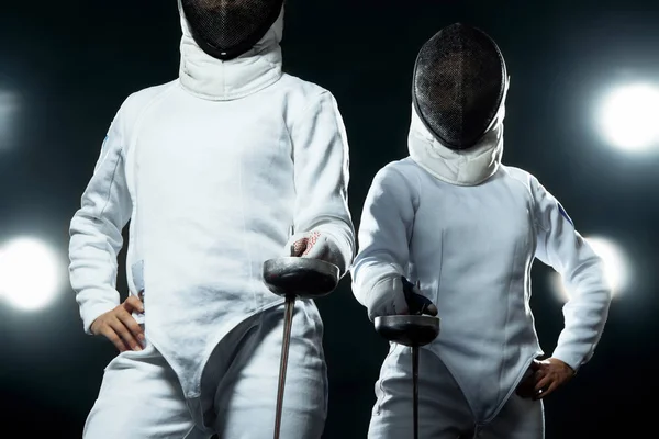 Young fencer athlete wearing mask and white fencing costume. holding the sword on black background with lights. — Stock Photo, Image