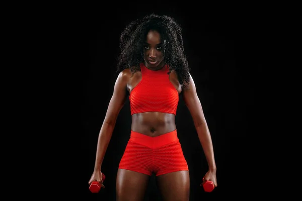 Deportiva mujer de piel negra en ropa deportiva roja, atleta con mancuernas hace ejercicio de fitness sobre fondo oscuro . — Foto de Stock