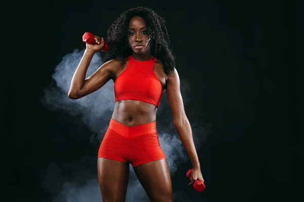 Mulher de pele preta ajuste desportivo em sportswear vermelho, atleta com halteres faz exercício fitness no fundo escuro . — Fotografia de Stock