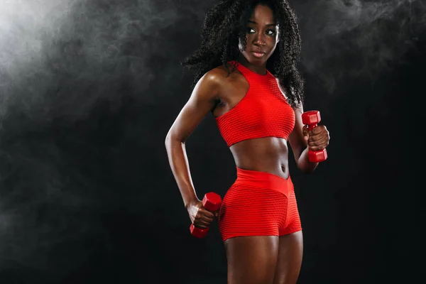 Mulher de pele preta ajuste desportivo em sportswear vermelho, atleta com halteres faz exercício fitness no fundo escuro . — Fotografia de Stock