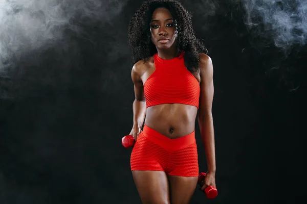 Mulher de pele preta ajuste desportivo em sportswear vermelho, atleta com halteres faz exercício fitness no fundo escuro . — Fotografia de Stock