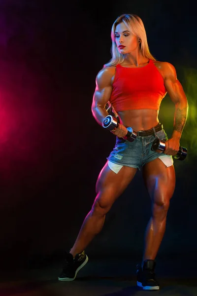Güçlü atletik kadın vücut geliştirmeci spor içinde giyen koyu arka plan üzerinde dumbbells ile. Fitness ve spor motivasyon. — Stok fotoğraf