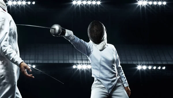 Two fencers on professional sports arena. Young athletes wearing mask and white fencing costume on duel in action. Women holding the sword on black background with lights. — Stock Photo, Image