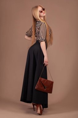 Moda kıyafetleri giyen, çantalı ve kahverengi arka planda güneş gözlüğü takan bir kadın..