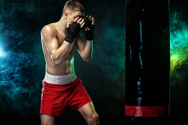 Deportista, hombre boxeador luchando con guantes con saco de boxeo. Aislado sobre fondo negro con humo. Copiar espacio . — Foto de Stock