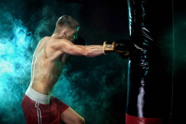 Esportista, boxeador lutando com luvas com saco de boxe. Isolado em fundo preto com fumaça. Espaço de cópia . — Fotografia de Stock