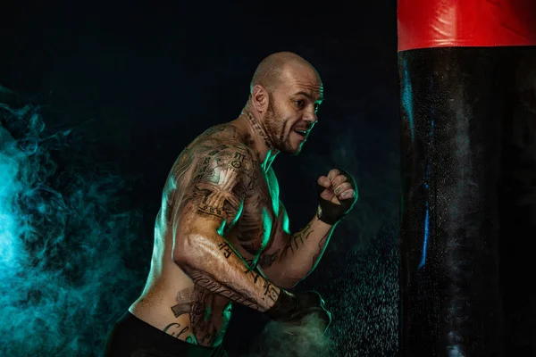 Deportista con tatuajes, boxeador hombre luchando en guantes con saco de boxeo. Aislado sobre fondo negro con humo. Copiar espacio . —  Fotos de Stock